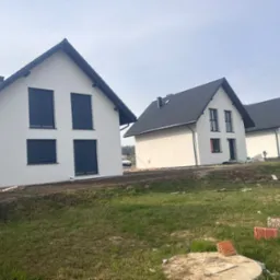 Trzy identyczne, nowo wybudowane domy z białymi elewacjami i czarnymi dachami, ustawione w rzędzie na trawiastym terenie. Widoczne cegły i elementy budowlane.