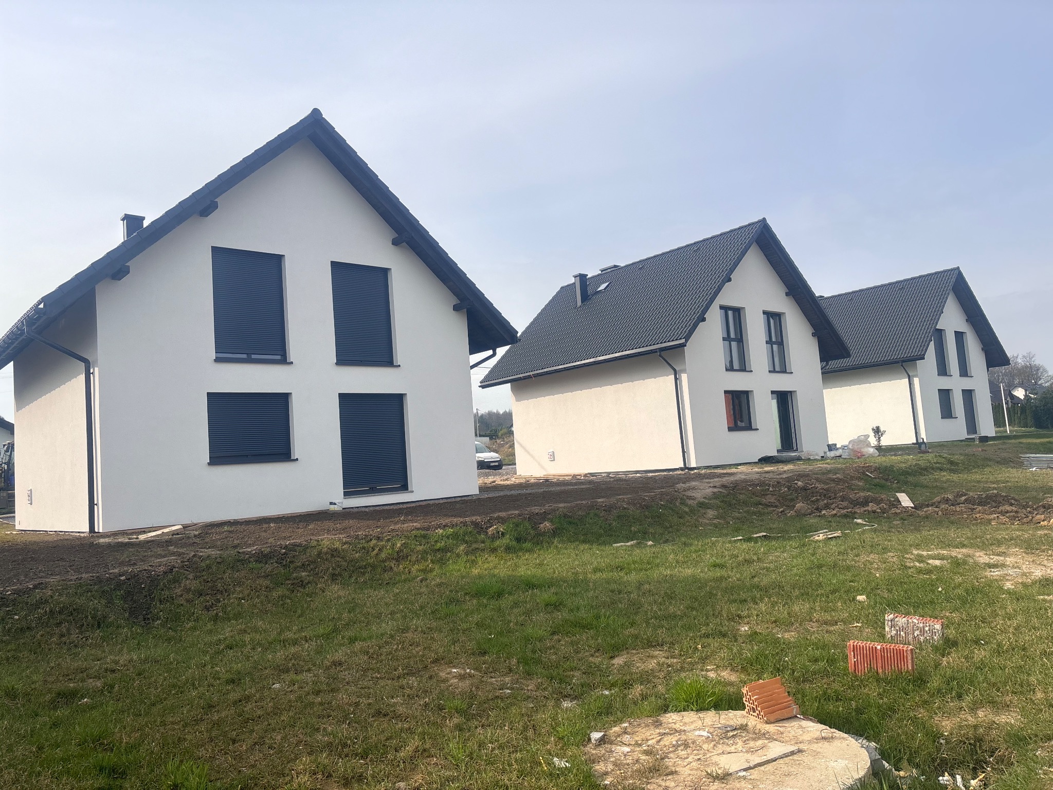 Trzy identyczne, nowo wybudowane domy z białymi elewacjami i czarnymi dachami, ustawione w rzędzie na trawiastym terenie. Widoczne cegły i elementy budowlane.