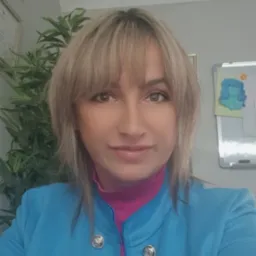 Portret uśmiechniętej kobiety w niebieskiej marynarce i różowym golfie, w tle roślina doniczkowa i tablica suchościeralna z rysunkiem.