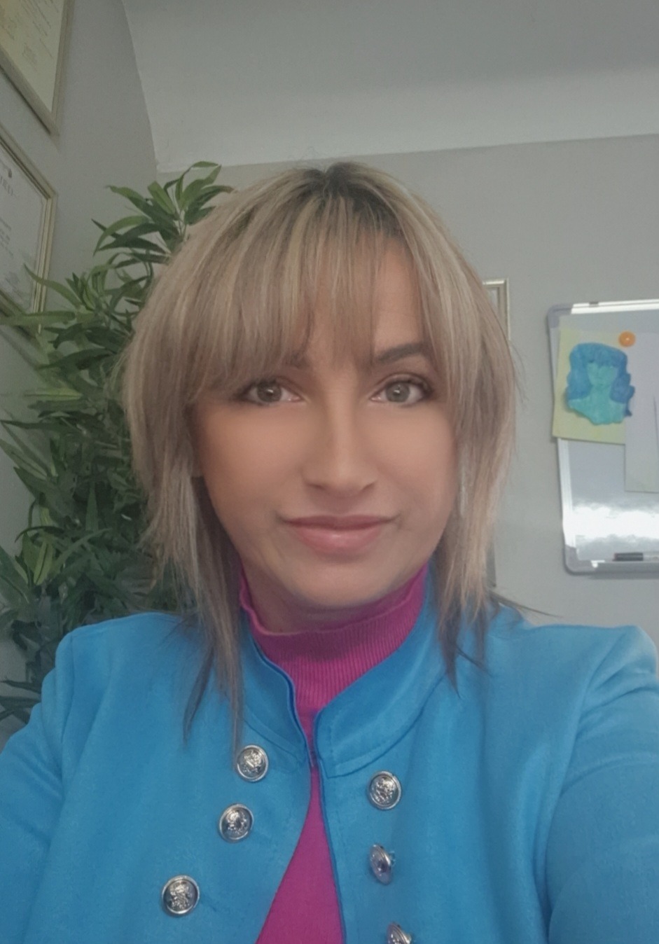 Portret uśmiechniętej kobiety w niebieskiej marynarce i różowym golfie, w tle roślina doniczkowa i tablica suchościeralna z rysunkiem.