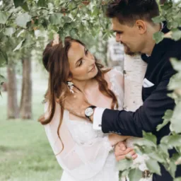 Para młoda w otoczeniu zieleni, panna młoda w zwiewnej sukni z koronkowymi detalami i delikatnymi kolczykami, pan młody w granatowym garniturze z eleganckim zegarkiem, oboje spoglądają na siebie...