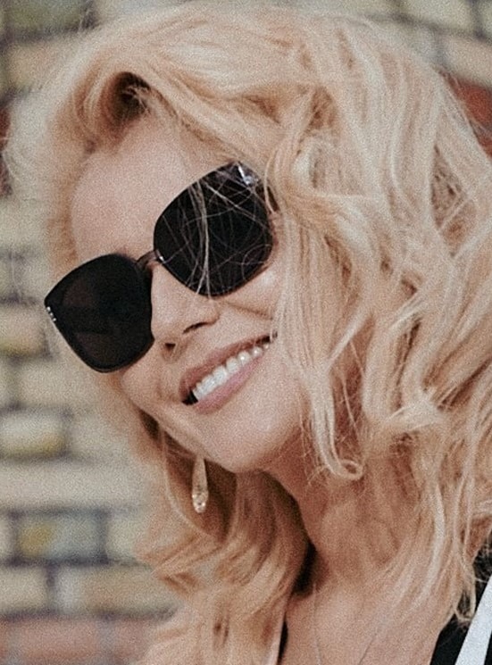 Uśmiechnięta blondynka w czarnych okularach przeciwsłonecznych i z wiszącymi kolczykami na tle ceglanej ściany.