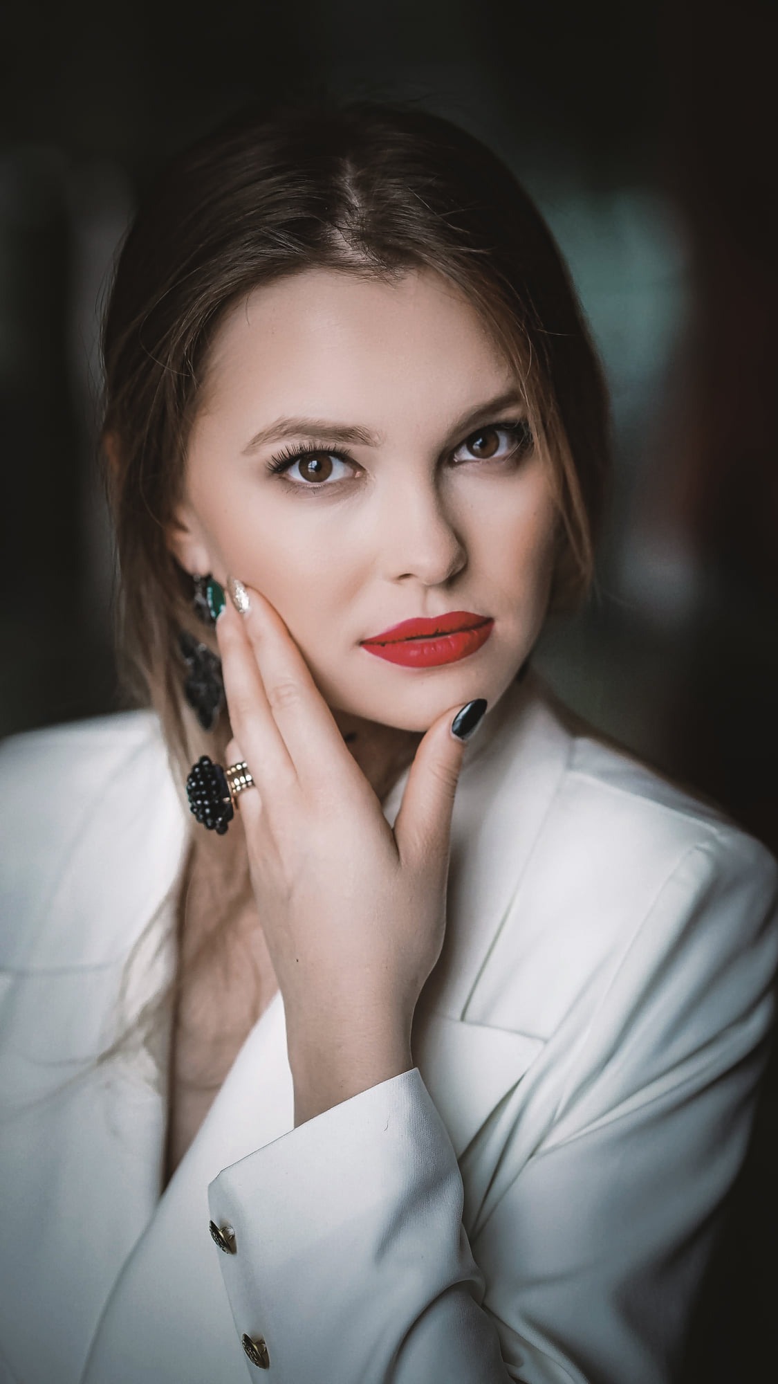 Portret kobiety w białym żakiecie, z wyrazistą czerwoną szminką, czarnym manicure, dużym pierścionkiem i kolczykami, dotykającej dłonią policzka.