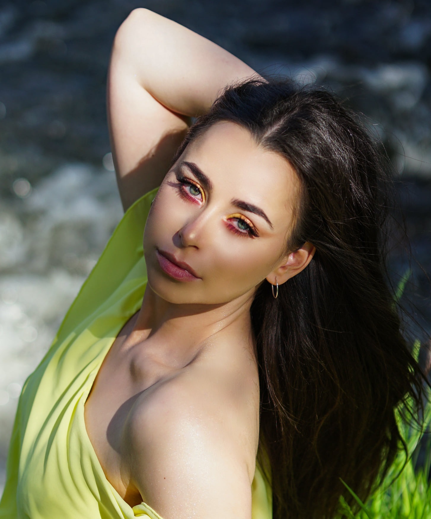 Portret kobiety z intensywnym makijażem oczu w odcieniach żółci i czerwieni, pozującej na tle rozmytej rzeki i zielonej trawy, ubrana w zwiewną, jasnożółtą tkaninę.