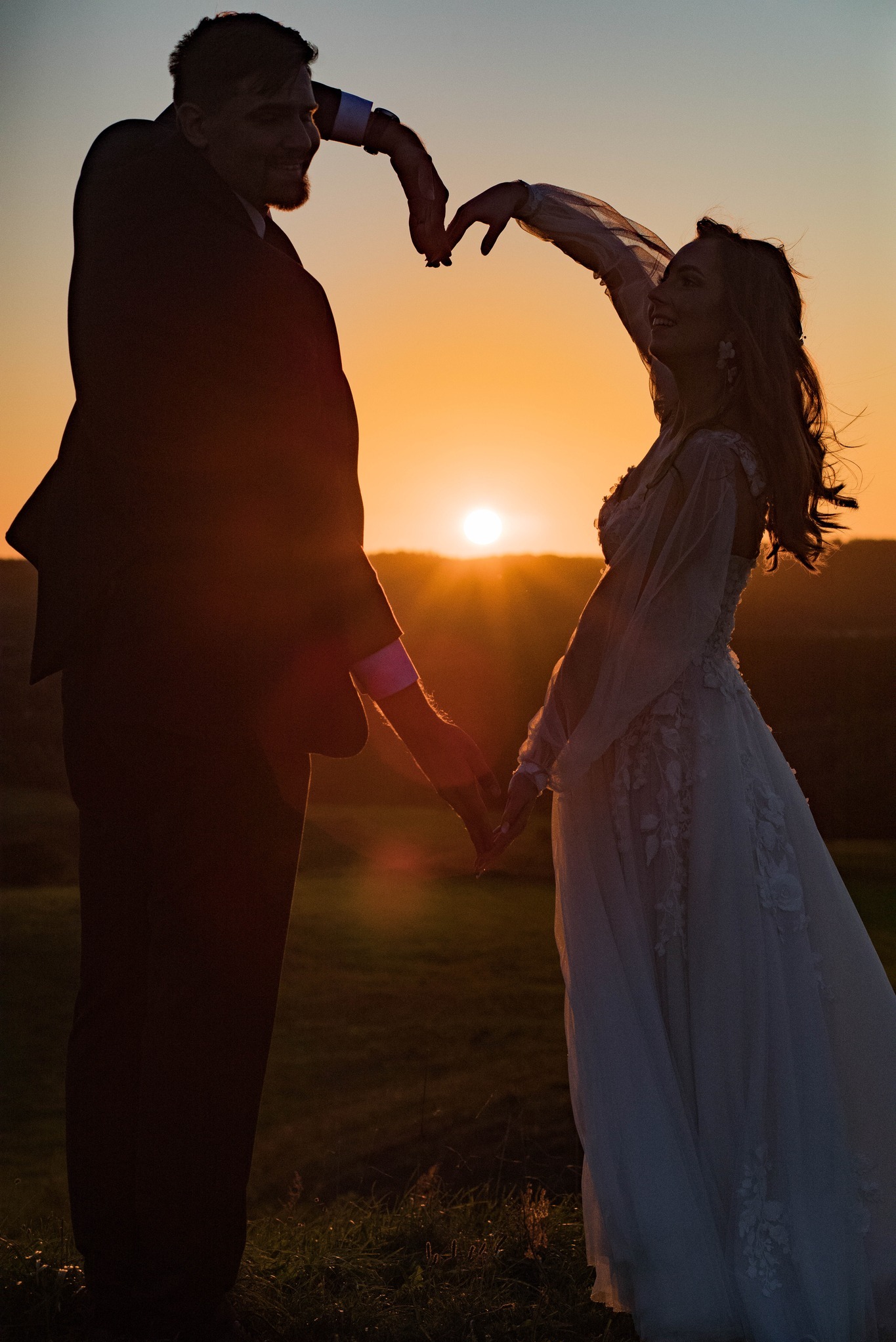 Silhouettes of a bride in a lace wedding dress and groom against a sunset, hands forming a heart shape.