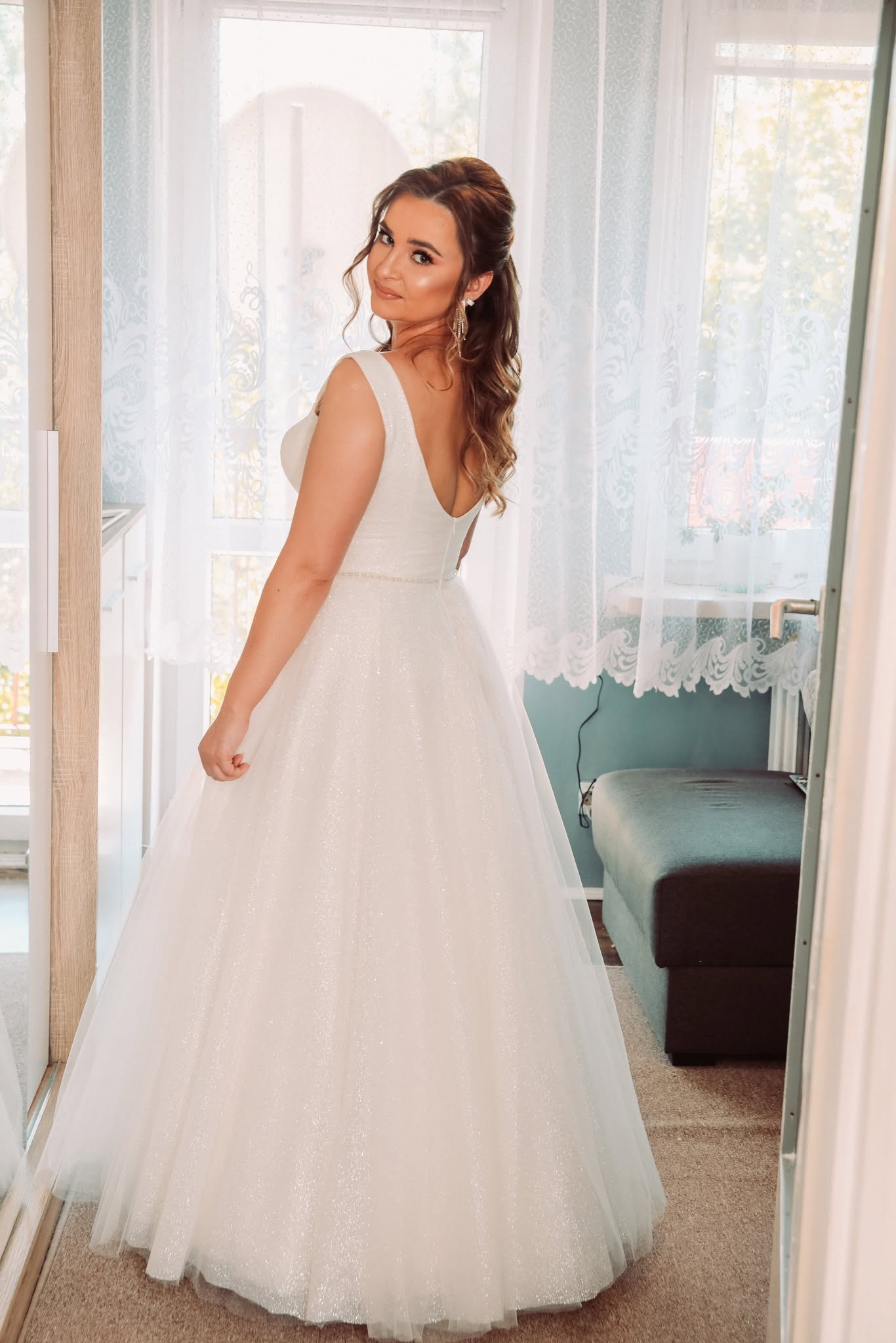 Uśmiechnięta panna młoda w białej, błyszczącej sukni balowej, spoglądająca przez ramię w lustrze, z widocznym fragmentem pokoju z oknem w tle.