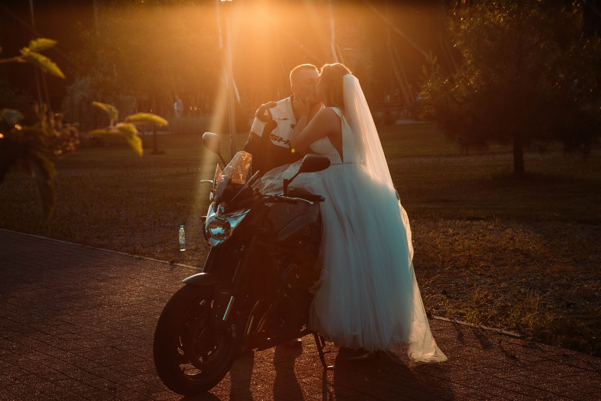 Para młoda w strojach ślubnych całuje się na motocyklu w promieniach zachodzącego słońca, widoczna butelka wody obok motocykla.