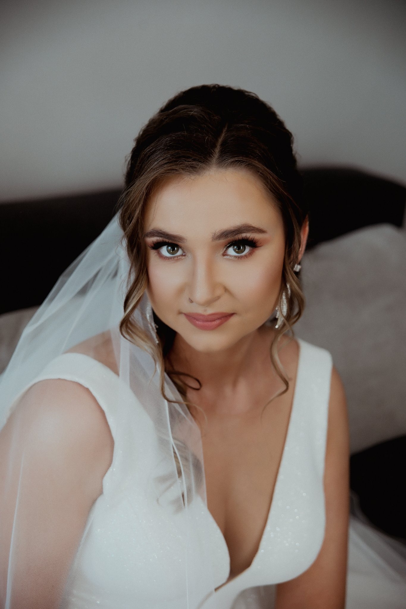 Portret panny młodej w białej sukni z welonem, delikatny makijaż i upięte włosy, zbliżenie na twarz i ramiona, spokojny wyraz twarzy, srebrne kolczyki.