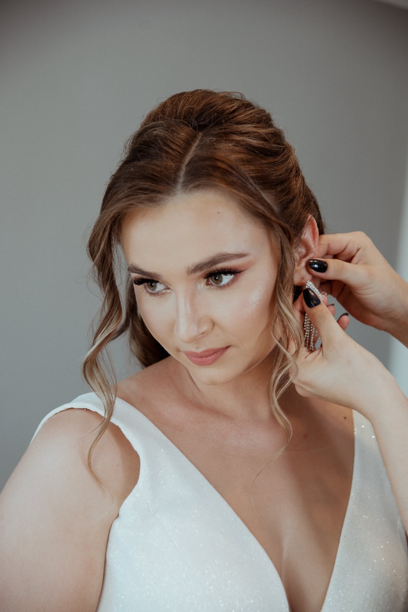 Panna młoda w białej sukni, z delikatnym makijażem, podczas zakładania błyszczących kolczyków przez wizażystkę z czarnym manicure.