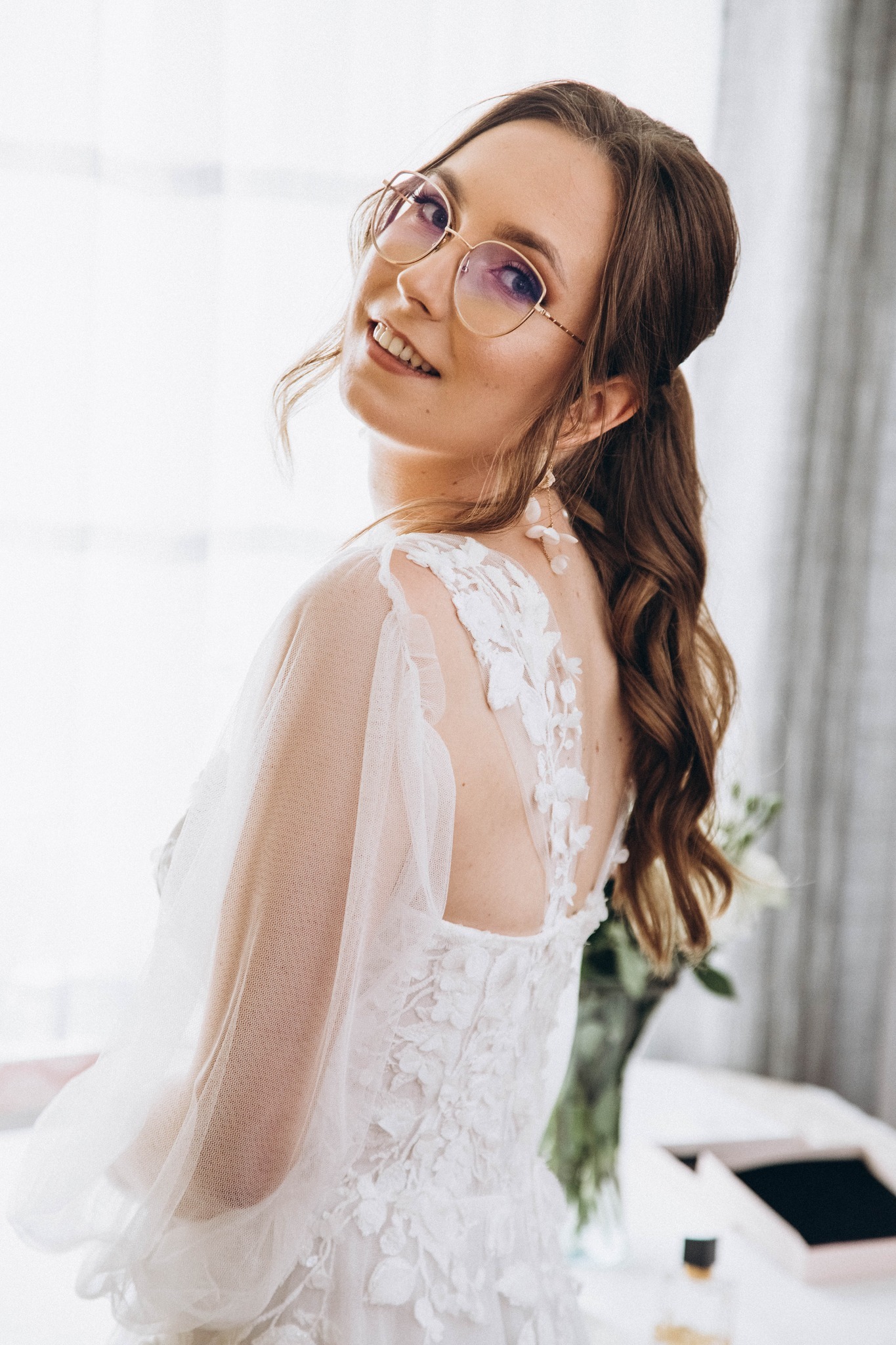 Uśmiechnięta panna młoda w okularach, z upiętymi włosami i koronkową suknią z delikatnymi rękawami z tiulu, spogląda przez ramię na jasnym tle okna.