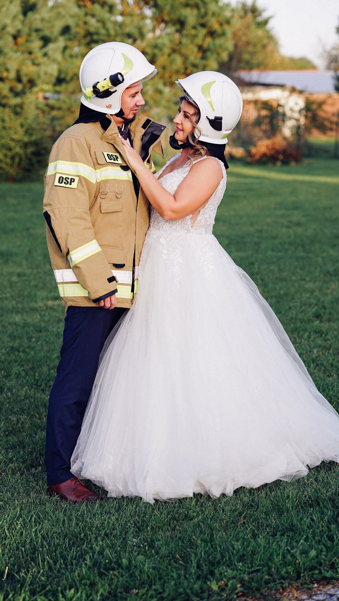 Para młoda w strojach ślubnych i hełmach strażackich, stojąca na trawie, panna młoda dotyka hełmu pana młodego, widoczne insygnia OSP na mundurze.