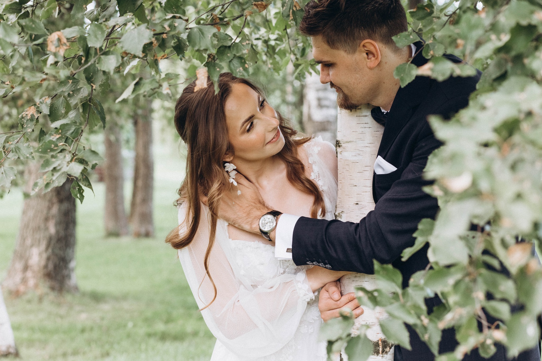 Para młoda w otoczeniu zieleni, panna młoda w zwiewnej sukni z koronkowymi detalami i delikatnymi kolczykami, pan młody w granatowym garniturze z eleganckim zegarkiem, oboje spoglądają na siebie...