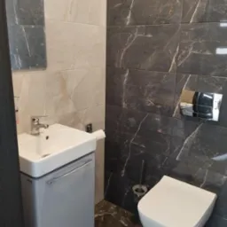 Nowoczesna toaleta z umywalką nablatową na szafce w łazience wykończonej płytkami imitującymi marmur w odcieniach beżu i grafitu, z widocznym lustrem i akcesoriami.