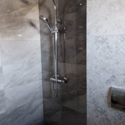 Klaczyński Zbigniew - Nowoczesna kabina prysznicowa z deszczownicą i słuchawką, wykończona płytkami imitującymi marmur w odcieniach szarości, z widocznym przyciskiem spłuczki w tle.