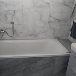 Klaczyński Zbigniew - Biała wanna obudowana szarymi płytkami imitującymi marmur, z baterią prysznicową na ścianie wyłożonej białymi płytkami marmuropodobnymi, w tle okno z dekoracjami.