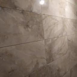 Ściana wyłożona beżowymi płytkami imitującymi marmur, z widocznymi krzyżykami dystansowymi i poziomnicą w dolnym rogu, wskazującymi na trwający proces układania.