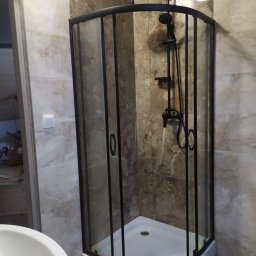 Klaczyński Zbigniew - Narożna kabina prysznicowa z czarną ramą i armaturą na tle marmurowych płytek w łazience.