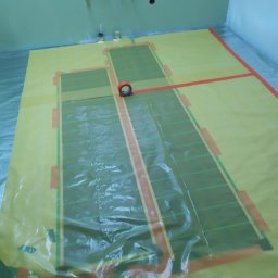 Klaczyński Zbigniew - Przygotowanie do montażu ogrzewania podłogowego w łazience, widoczna folia ochronna na podłodze z naniesionym schematem rozmieszczenia mat grzewczych i rolka taśmy klejącej.