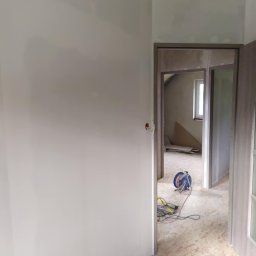 Klaczyński Zbigniew - Świeżo pomalowana ściana w jasnym odcieniu szarości, widoczna instalacja elektryczna i częściowo wykończone poddasze w tle, z rozłożonymi materiałami budowlanymi i żółtą wełną mineralną.