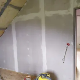 Poddasze w trakcie wykańczania: ocieplenie wełną mineralną, płyty gipsowo-kartonowe z widocznymi łączeniami szpachlowanymi, niezabezpieczone przewody elektryczne wystające ze ściany, oraz wiadro...