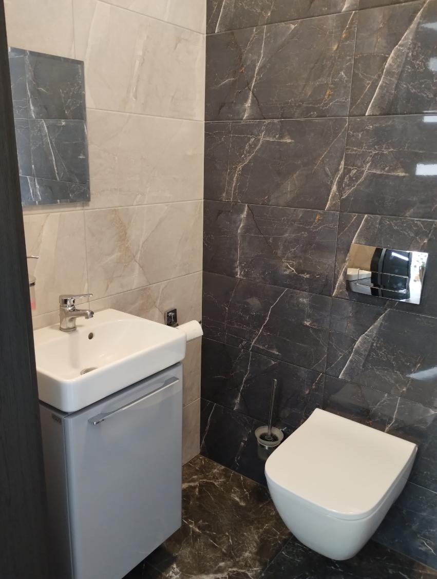 Nowoczesna toaleta z umywalką nablatową na szafce w łazience wykończonej płytkami imitującymi marmur w odcieniach beżu i grafitu, z widocznym lustrem i akcesoriami.