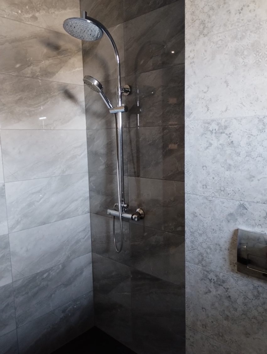 Nowoczesna kabina prysznicowa z deszczownicą i słuchawką, wykończona płytkami imitującymi marmur w odcieniach szarości, z widocznym przyciskiem spłuczki w tle.