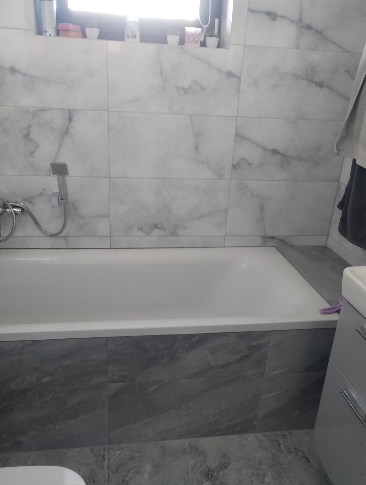 Biała wanna obudowana szarymi płytkami imitującymi marmur, z baterią prysznicową na ścianie wyłożonej białymi płytkami marmuropodobnymi, w tle okno z dekoracjami.