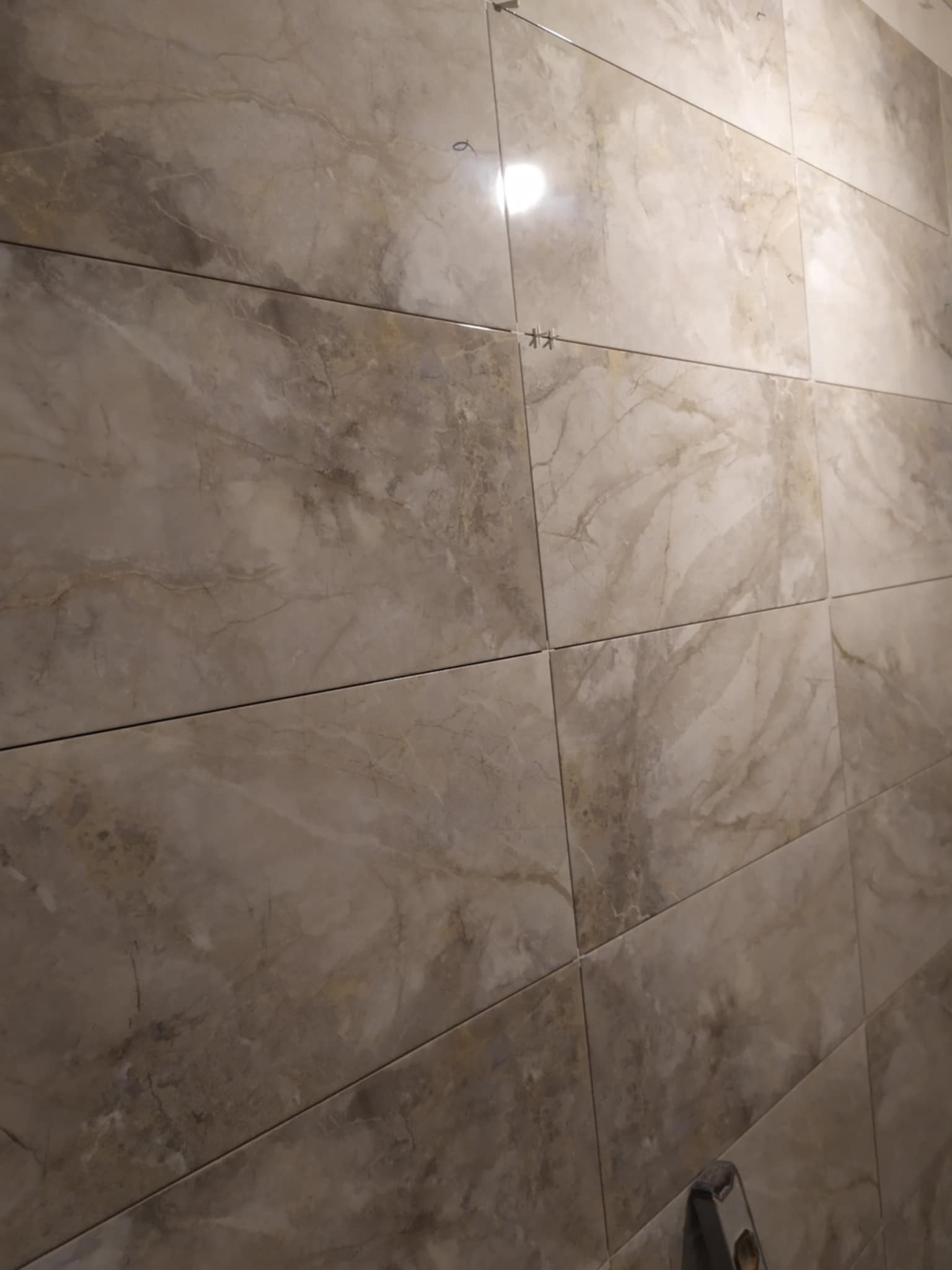 Ściana wyłożona beżowymi płytkami imitującymi marmur, z widocznymi krzyżykami dystansowymi i poziomnicą w dolnym rogu, wskazującymi na trwający proces układania.