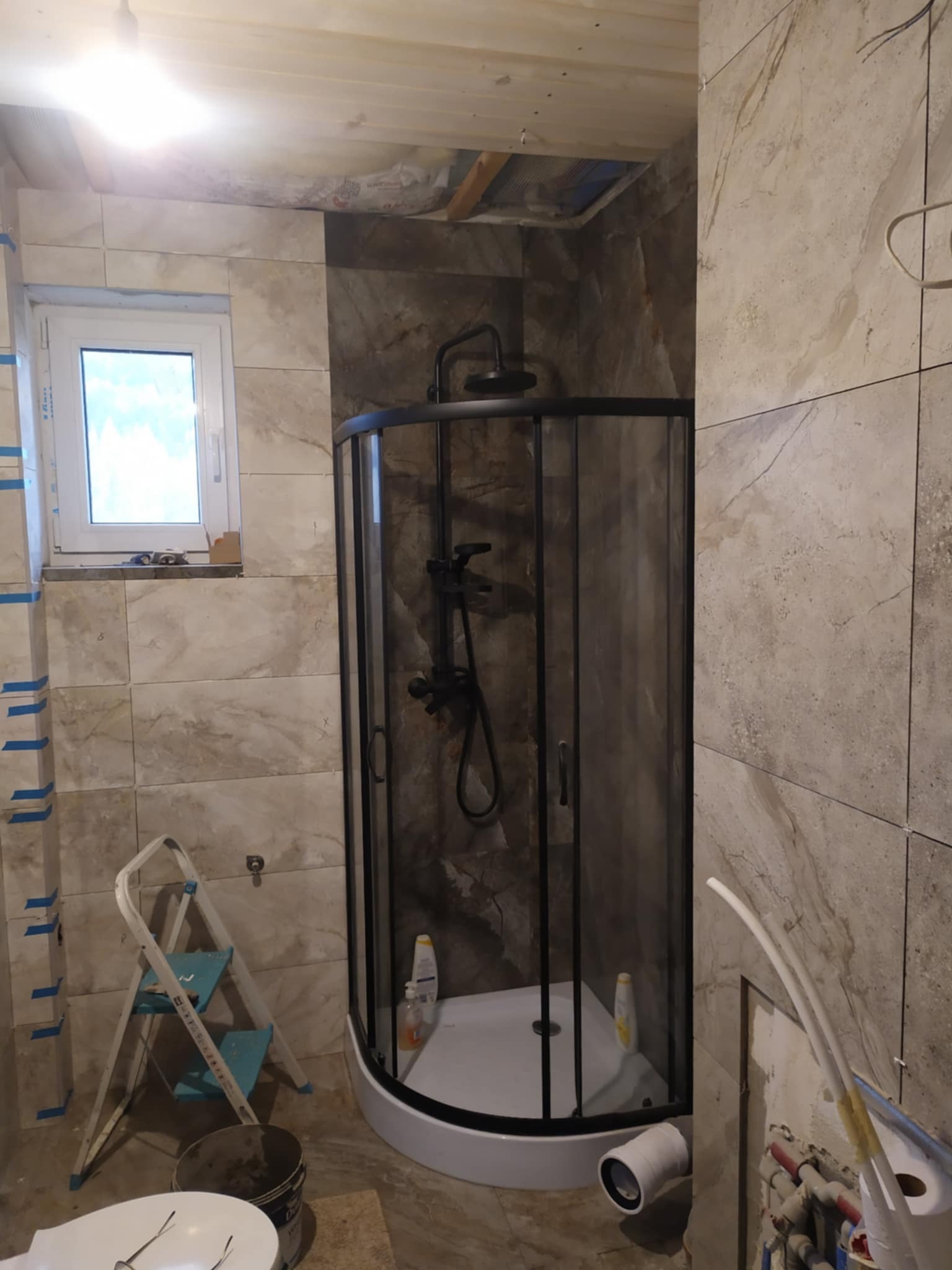 Niedokończona łazienka z kabiną prysznicową w czarnej ramie, widoczne narzędzia i materiały budowlane, płytki imitujące marmur na ścianach, sufit częściowo odkryty.