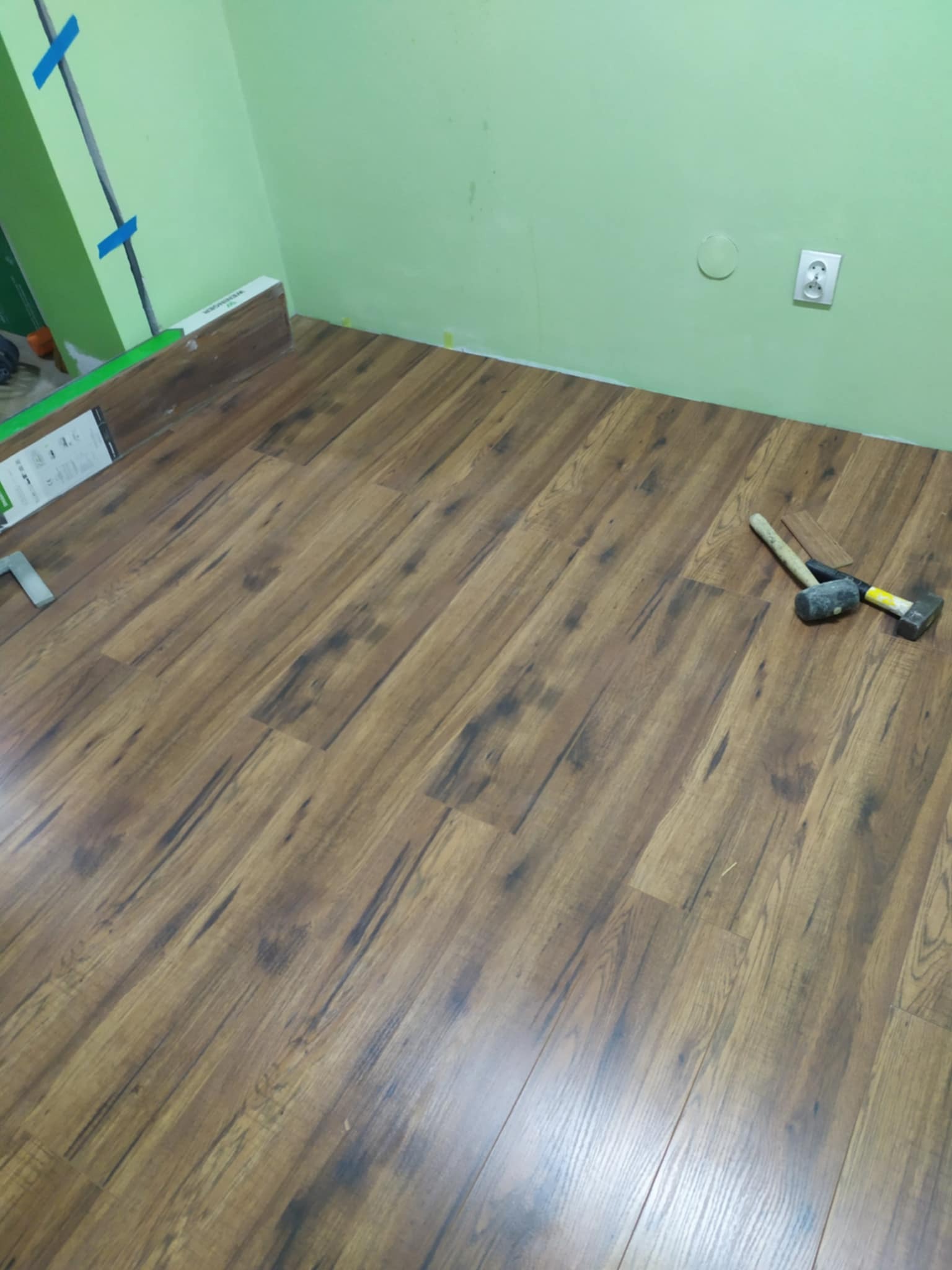 Ułożona podłoga z paneli imitujących drewno w jasnym odcieniu brązu, widoczne dwa młotki i resztki paneli przy ścianie, zielona ściana z gniazdkiem elektrycznym.