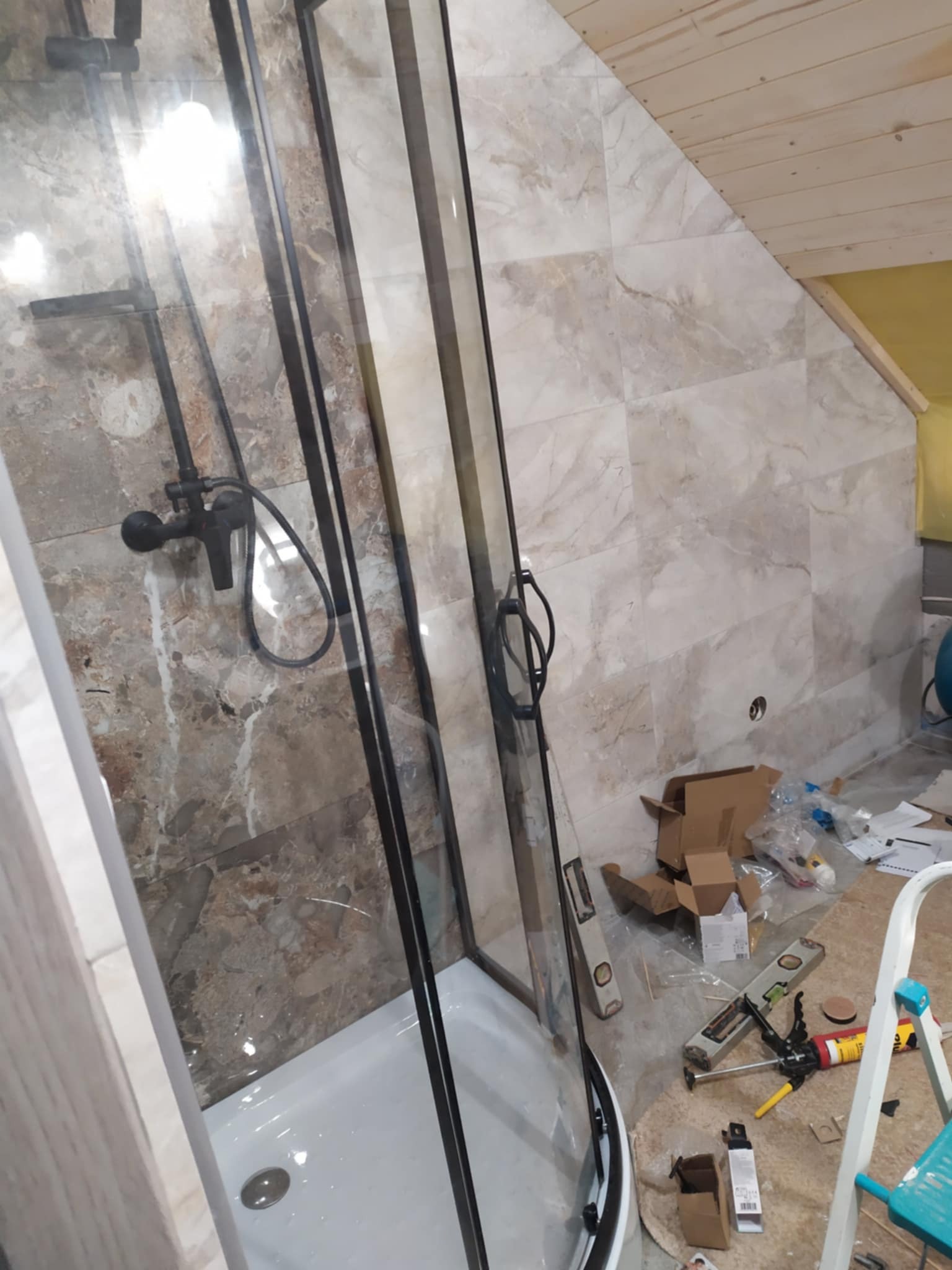 Montaż kabiny prysznicowej z czarnymi profilami w łazience z marmurowymi płytkami, widoczne narzędzia i drabina.