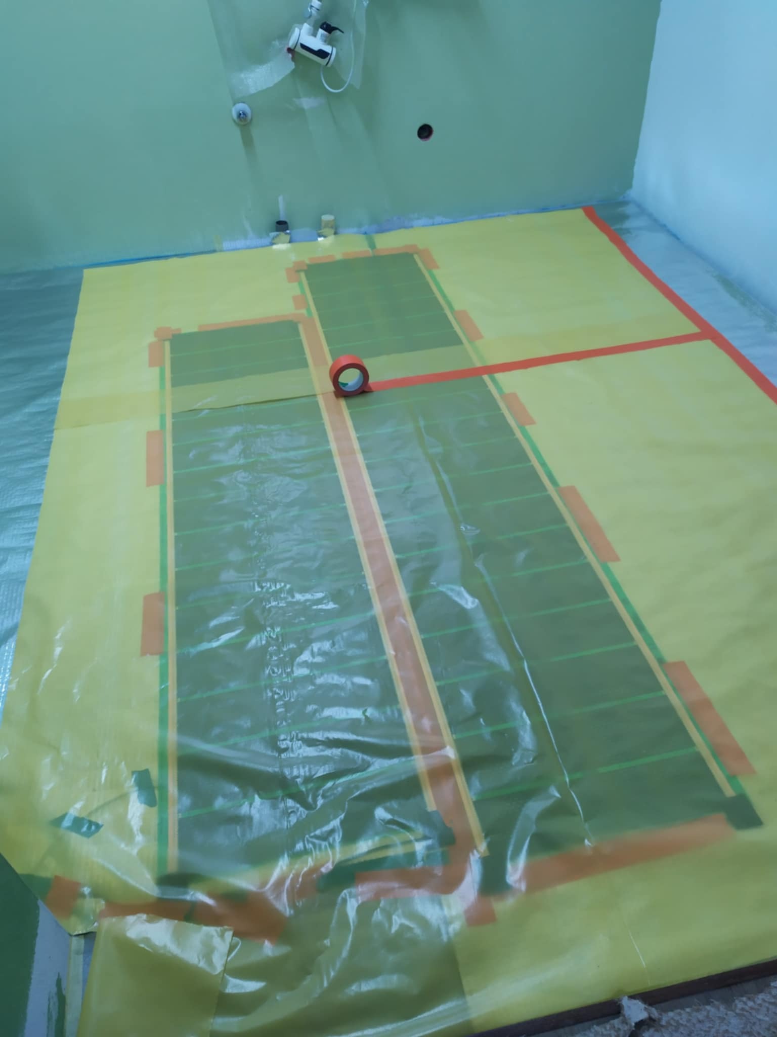 Przygotowanie do montażu ogrzewania podłogowego w łazience, widoczna folia ochronna na podłodze z naniesionym schematem rozmieszczenia mat grzewczych i rolka taśmy klejącej.