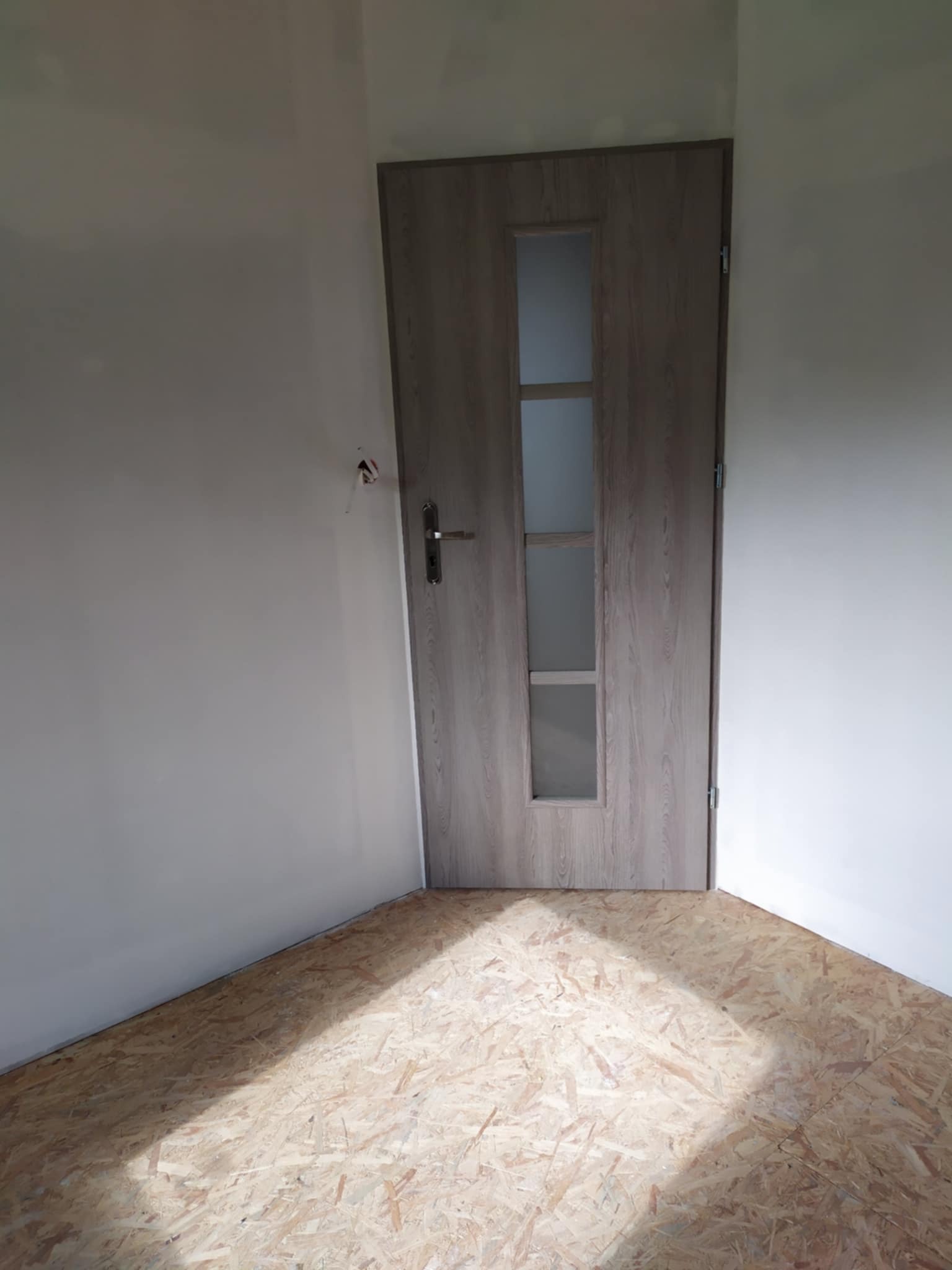 Wnętrze pokoju z zamontowanymi drzwiami wewnętrznymi z szybami, surowe ściany pomalowane na biało i podłoga wyłożona płytami OSB.