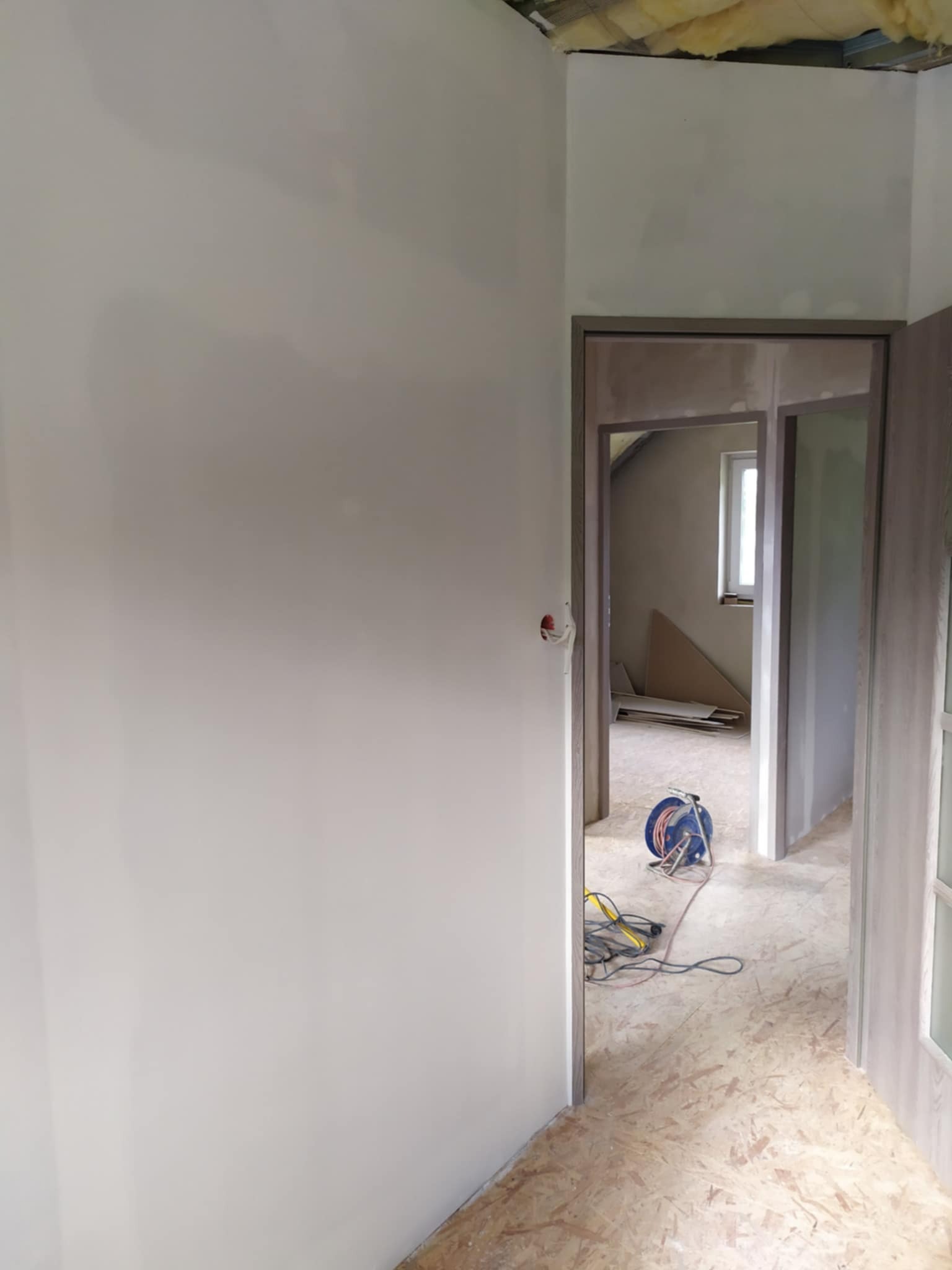 Świeżo pomalowana ściana w jasnym odcieniu szarości, widoczna instalacja elektryczna i częściowo wykończone poddasze w tle, z rozłożonymi materiałami budowlanymi i żółtą wełną mineralną.