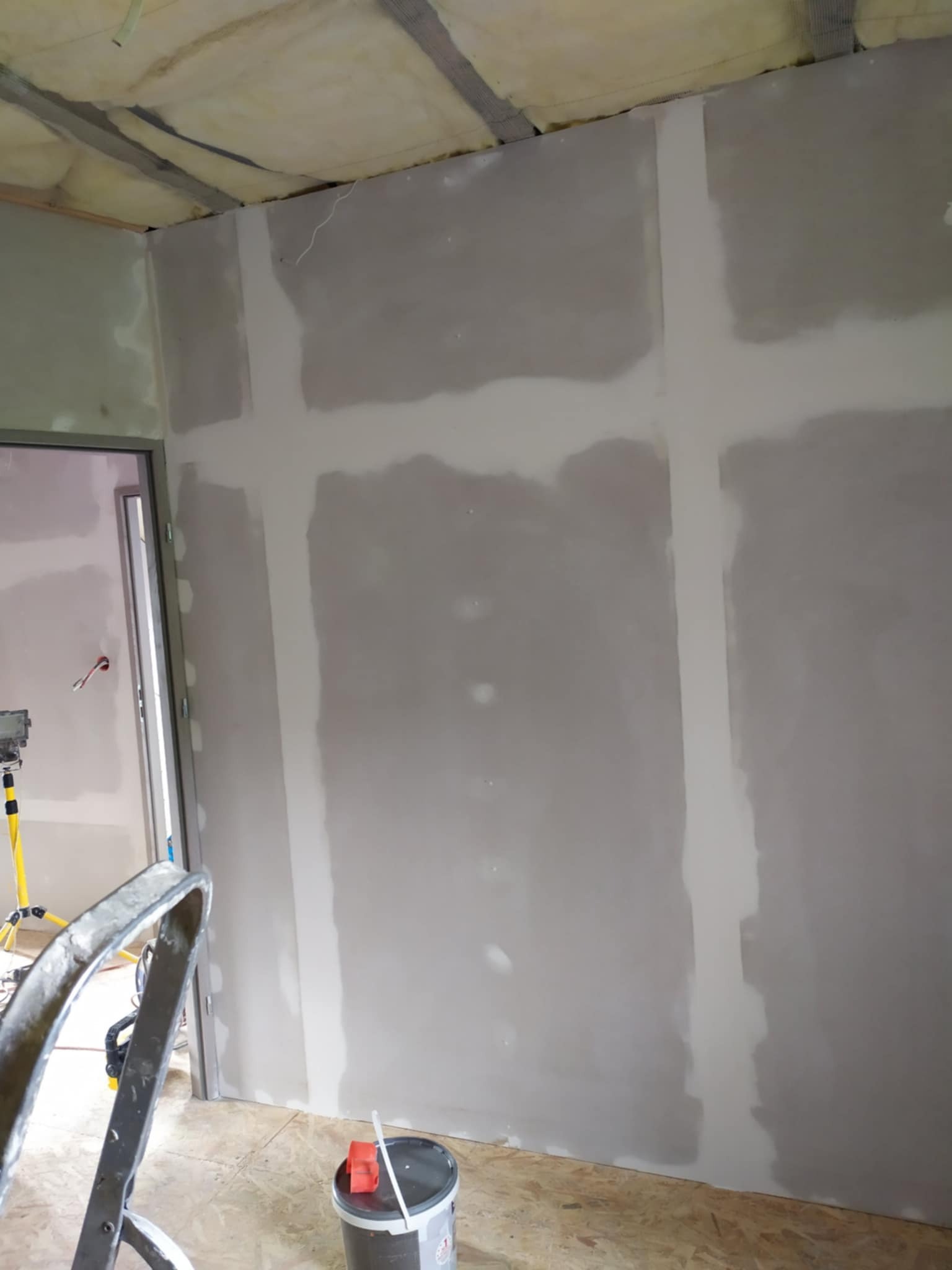 Wnętrze w trakcie szpachlowania płyt gipsowo-kartonowych, widoczne łączenia wzmocnione taśmą, drabina malarska i wiadro z masą szpachlową.