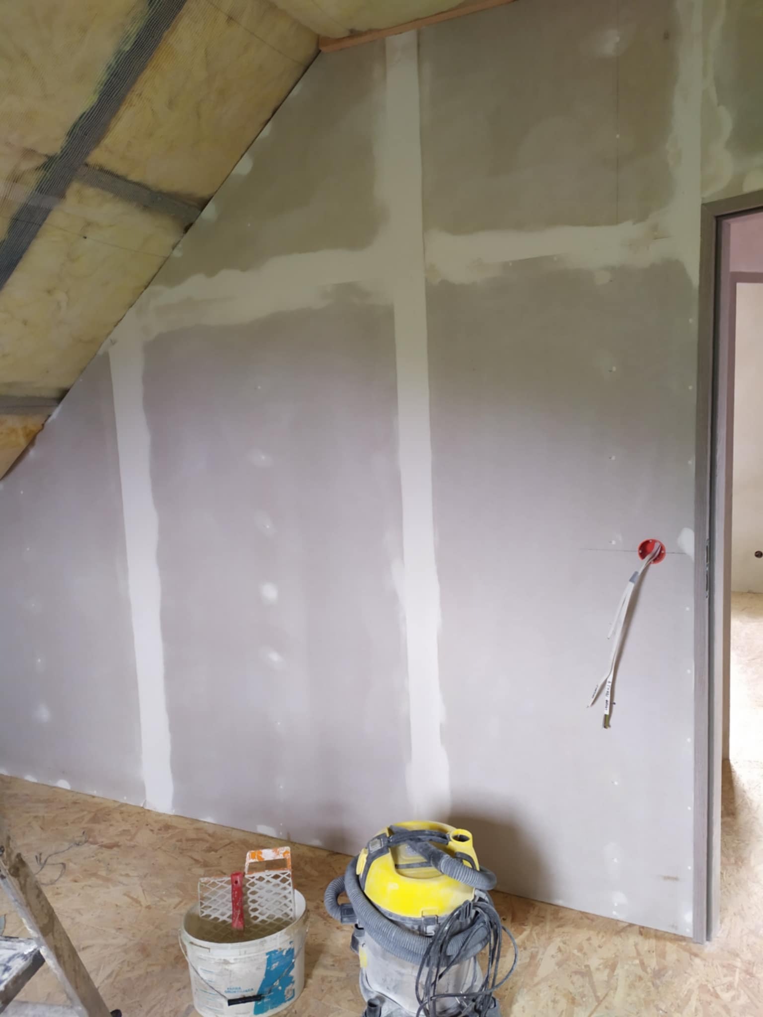 Poddasze w trakcie wykańczania: ocieplenie wełną mineralną, płyty gipsowo-kartonowe z widocznymi łączeniami szpachlowanymi, niezabezpieczone przewody elektryczne wystające ze ściany, oraz wiadro...