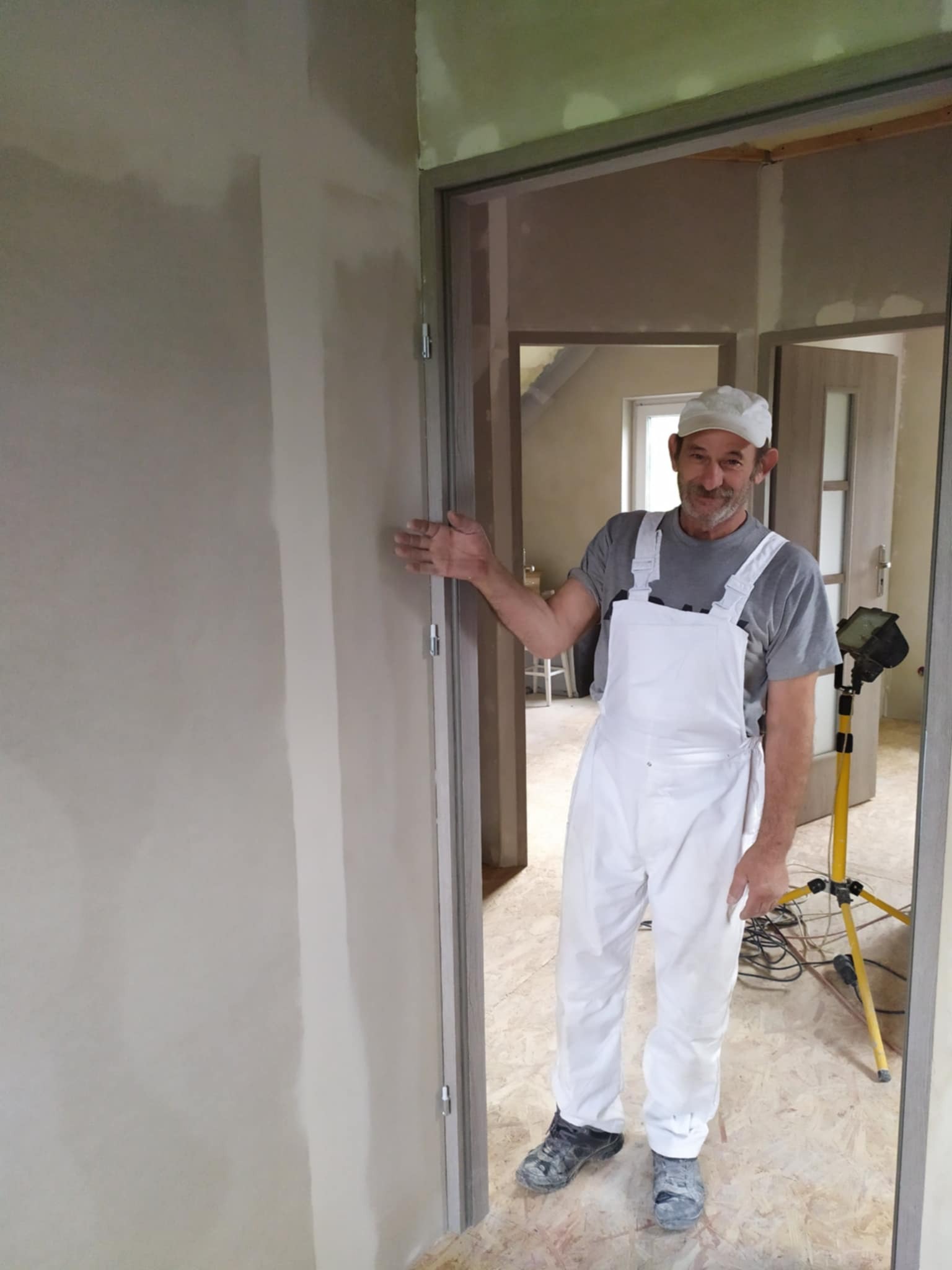 Mężczyzna w białym kombinezonie malarskim prezentuje świeżo zagruntowaną ścianę w trakcie remontu, widoczny reflektor na statywie i surowe wykończenie wnętrza.