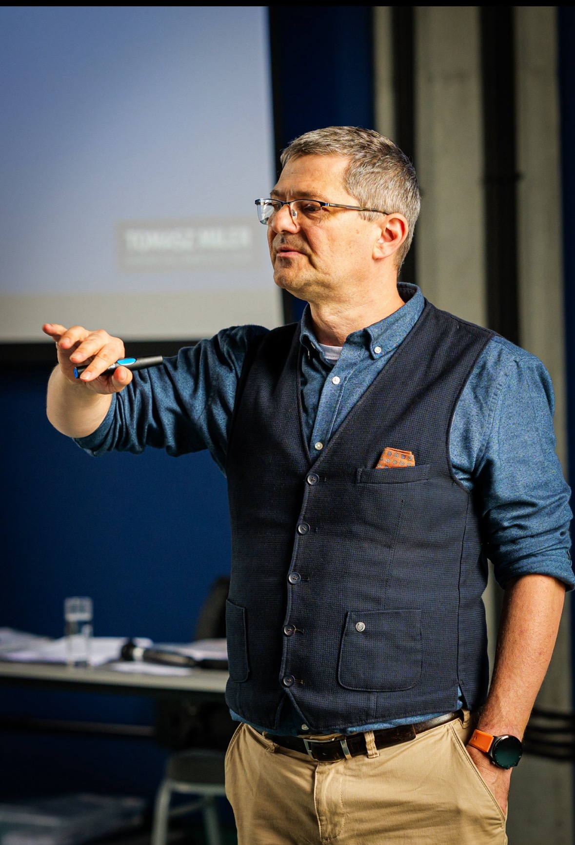 Mężczyzna w okularach, koszuli i kamizelce gestykuluje podczas prezentacji. W tle ekran projekcyjny i stół z dokumentami. Widoczny zegarek na ręce.