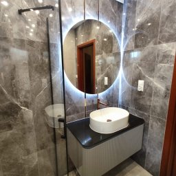 TOPFLIZ Roman Szarek - Nowoczesna łazienka z marmurowymi płytkami, okrągłym lustrem z podświetleniem LED, białą umywalką nablatową i szklaną kabiną prysznicową.