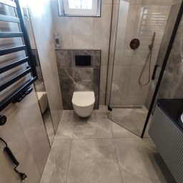 TOPFLIZ Roman Szarek - Nowoczesna łazienka z marmurowymi płytkami, wiszącą toaletą i kabiną prysznicową ze złotymi akcentami. Minimalistyczny design z grzejnikiem drabinkowym.