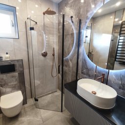 TOPFLIZ Roman Szarek - Elegancka łazienka w szarościach z kabiną prysznicową typu walk-in, umywalką nablatową i podświetlanym lustrem. Armatura w kolorze miedzi dodaje ciepła.