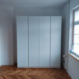 TOPFLIZ Roman Szarek - Biała, czterodrzwiowa szafa wnękowa na parkiecie ułożonym w jodełkę. Widok na fragment okna z butelką wody. Minimalistyczny design, jasne wnętrze.