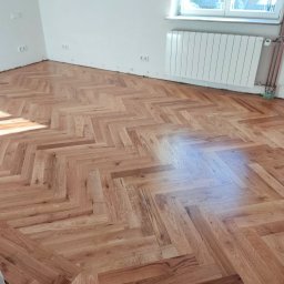 TOPFLIZ Roman Szarek - Ukończona podłoga z parkietu ułożonego w jodełkę, widoczna w jasnym, pustym pokoju z grzejnikiem i gniazdkami elektrycznymi na ścianie.