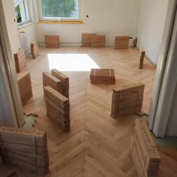 TOPFLIZ Roman Szarek - Układanie parkietu w jodełkę w jasnym pokoju z otwartym oknem. Pakiety desek ułożone na podłodze, częściowo pokrytej parkietem. Widoczne niedokończone fragmenty podłogi.