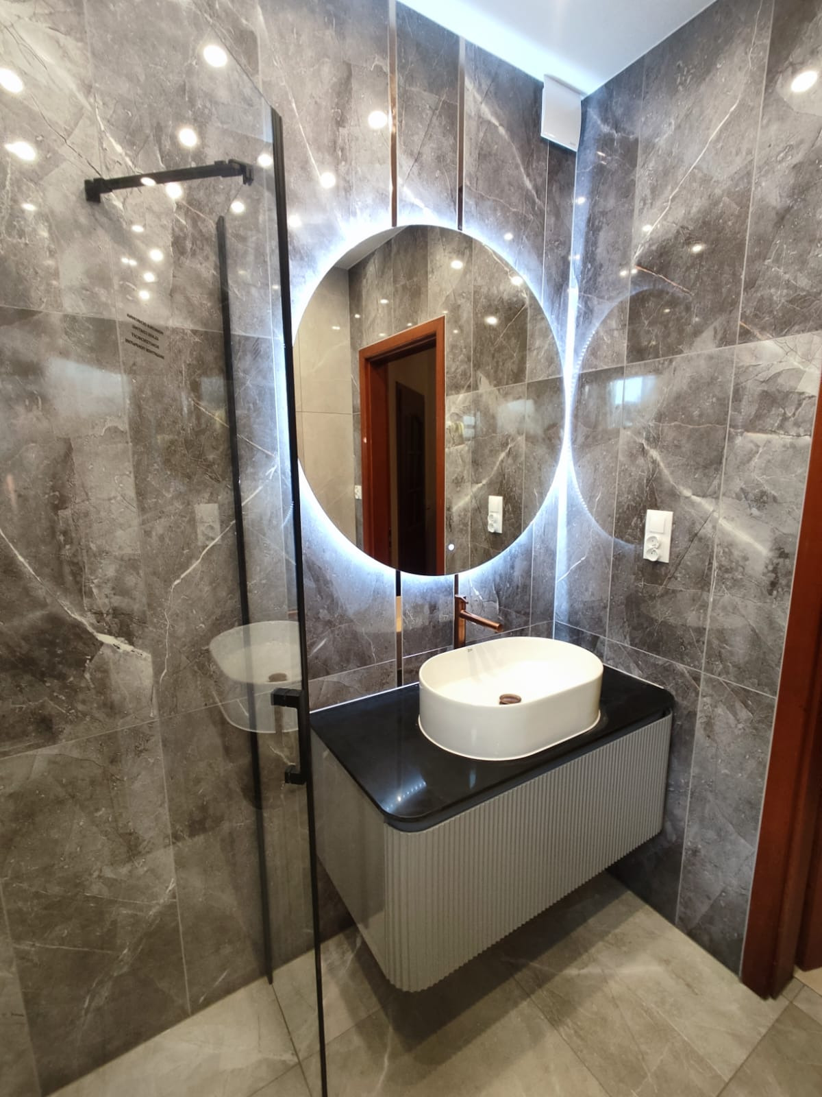 Nowoczesna łazienka z marmurowymi płytkami, okrągłym lustrem z podświetleniem LED, białą umywalką nablatową i szklaną kabiną prysznicową.