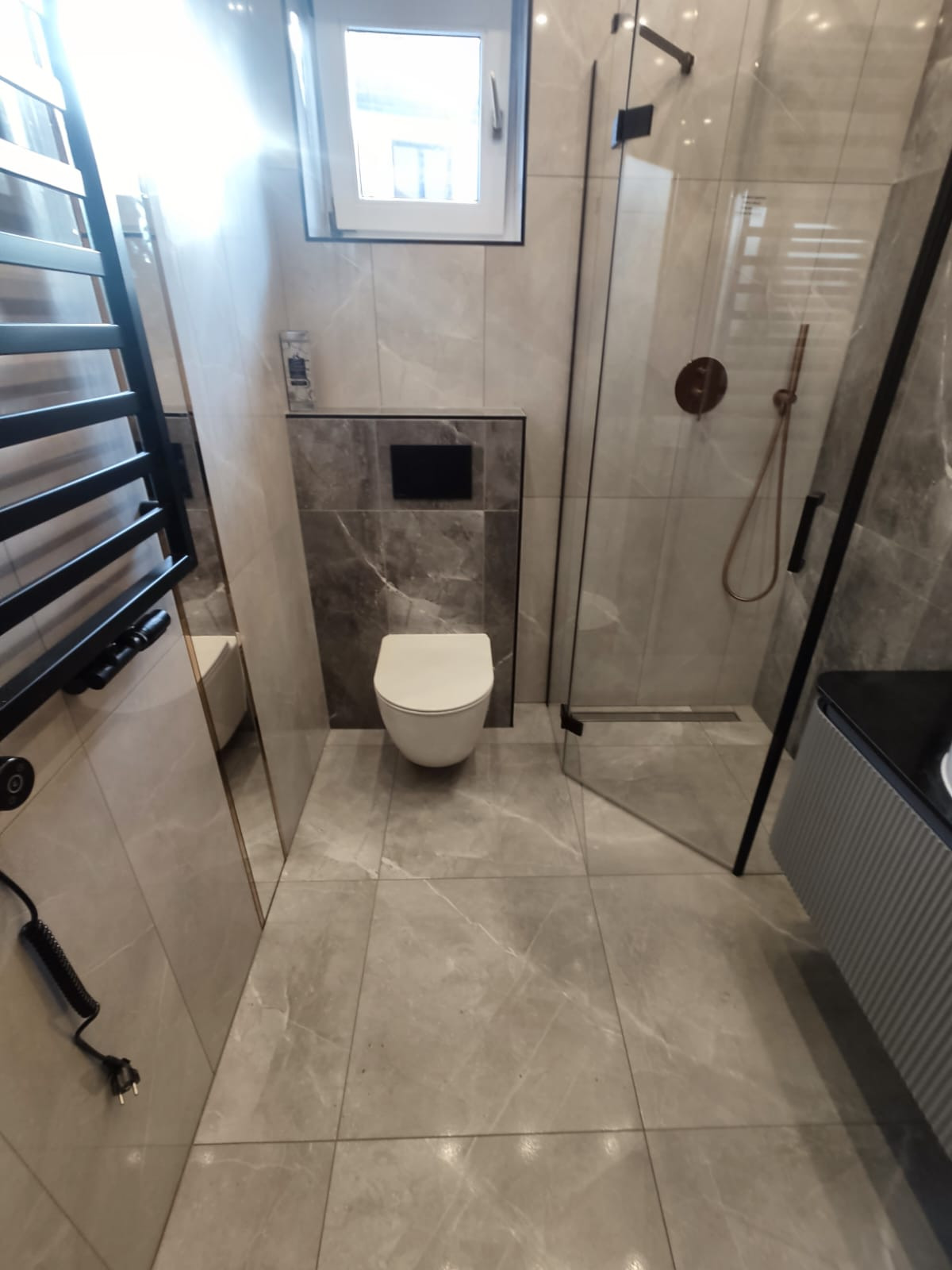 Nowoczesna łazienka z marmurowymi płytkami, wiszącą toaletą i kabiną prysznicową ze złotymi akcentami. Minimalistyczny design z grzejnikiem drabinkowym.