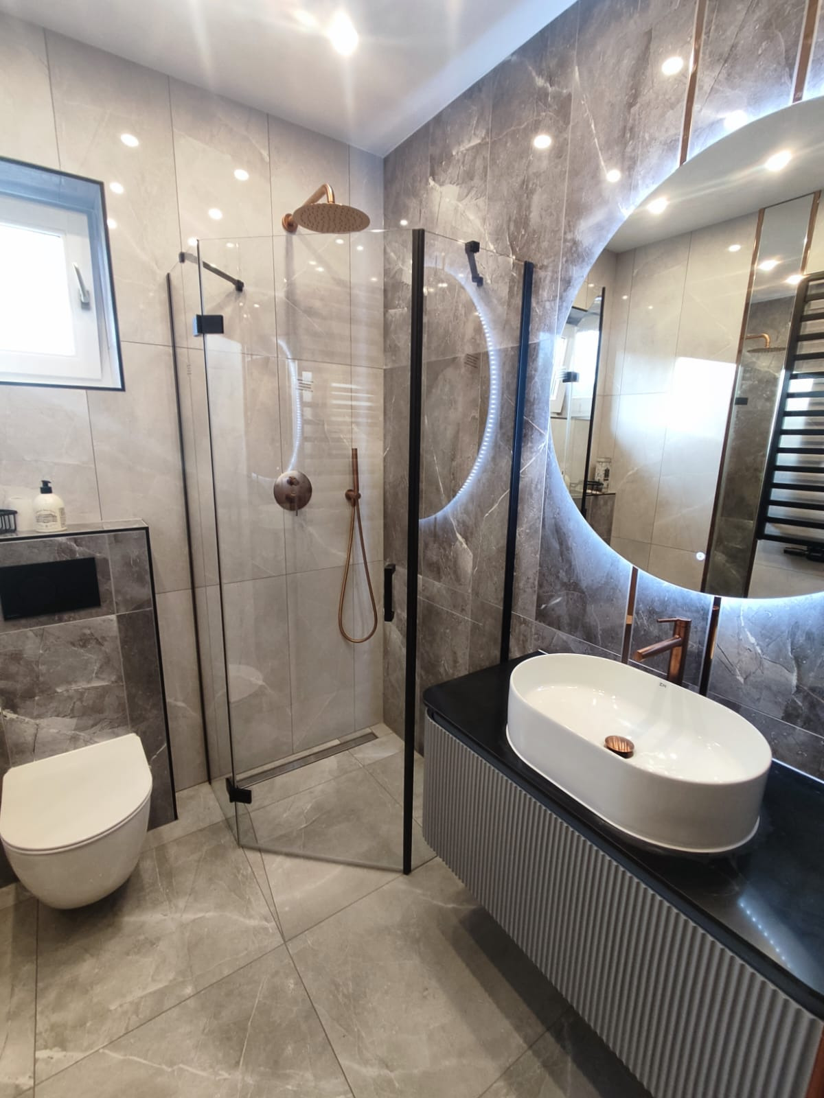 Elegancka łazienka w szarościach z kabiną prysznicową typu walk-in, umywalką nablatową i podświetlanym lustrem. Armatura w kolorze miedzi dodaje ciepła.