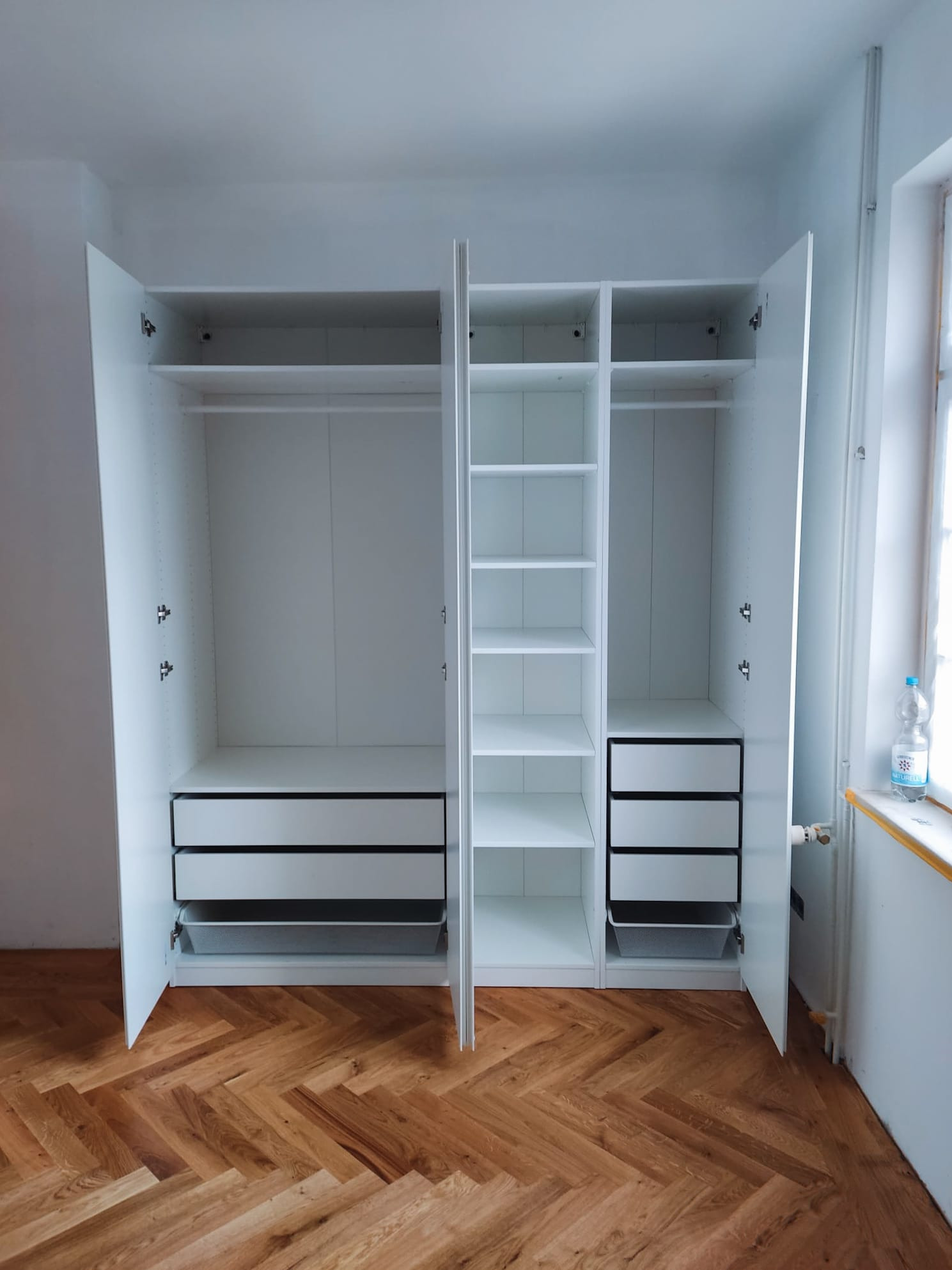 Biała, trzydrzwiowa szafa z szufladami i półkami, z otwartymi drzwiami, na parkiecie ułożonym w jodełkę. Widoczna butelka wody na parapecie.
