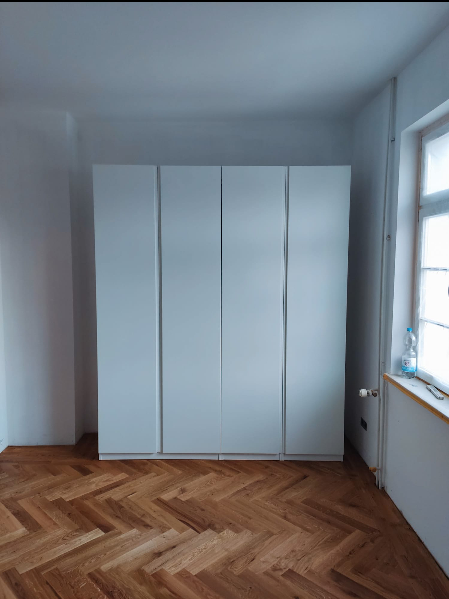 Biała, czterodrzwiowa szafa wnękowa na parkiecie ułożonym w jodełkę. Widok na fragment okna z butelką wody. Minimalistyczny design, jasne wnętrze.