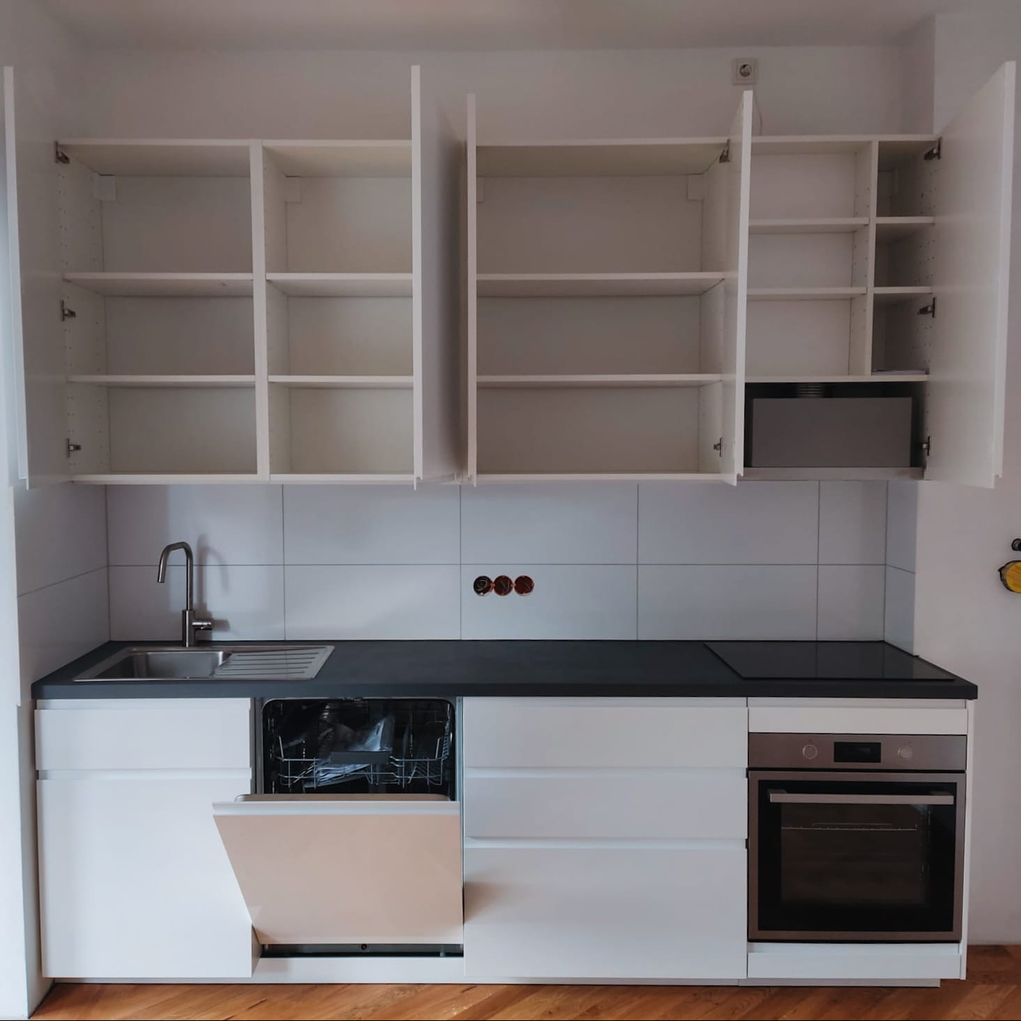 Biała, nowoczesna kuchnia z otwartymi szafkami wiszącymi, czarnym blatem i sprzętem AGD: piekarnik, płyta indukcyjna, zlew i otwarta zmywarka. Widoczne płytki na ścianie.